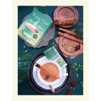Flat lay of Royal Elaichi (Cardamom) Unsweetened Chai box and sachet with a brewed cup of tea garnished with a cinnamon stick and star anise on a dark green background.