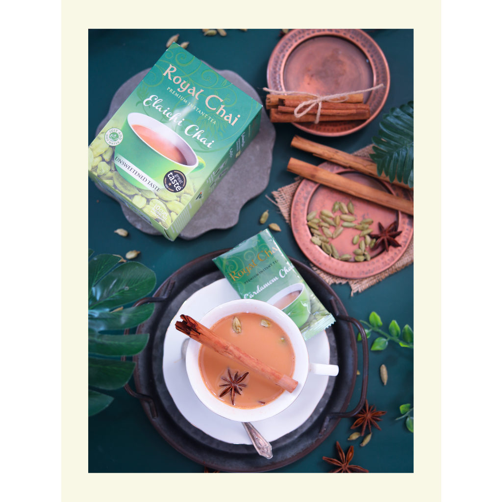 Flat lay of Royal Elaichi (Cardamom) Unsweetened Chai box and sachet with a brewed cup of tea garnished with a cinnamon stick and star anise on a dark green background.