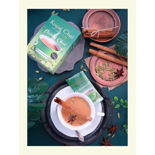 Flat lay of Royal Elaichi (Cardamom) Unsweetened Chai box and sachet with a brewed cup of tea garnished with a cinnamon stick and star anise on a dark green background.