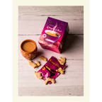 A box of Royal Unsweetened Ginger Chai on a rustic white wood surface, shown with a single serving sachet, fresh dried ginger pieces, and a traditional clay tea cup.