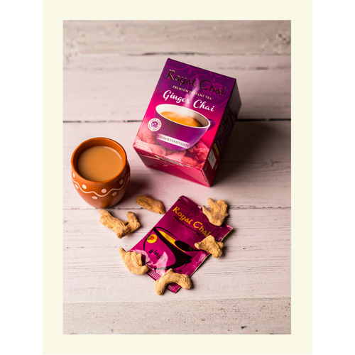 A box of Royal Unsweetened Ginger Chai on a rustic white wood surface, shown with a single serving sachet, fresh dried ginger pieces, and a traditional clay tea cup.
