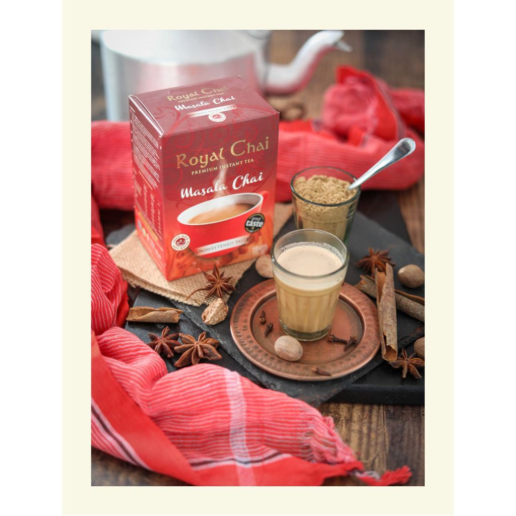 Royal Unsweetened Masala Chai box on a rustic wooden table with a glass of brewed tea, whole spices like star anise and nutmeg, and a traditional red cloth.